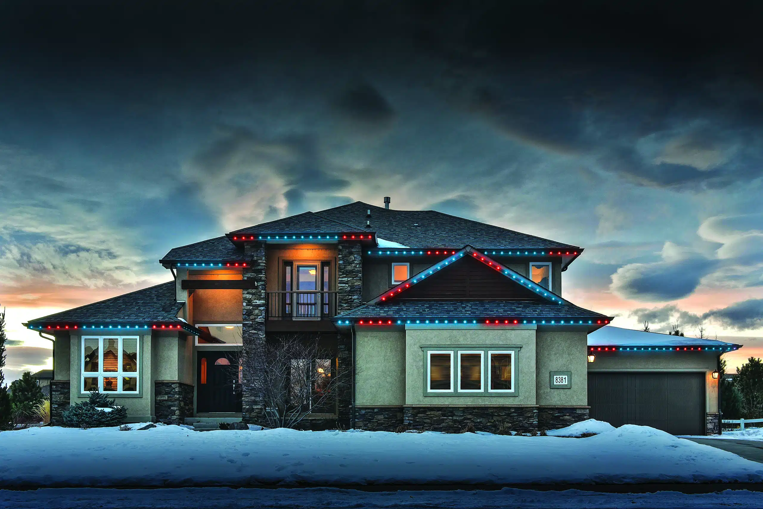 Sun setting behind winter home with permanent holiday lights glowing with snow filled front yard